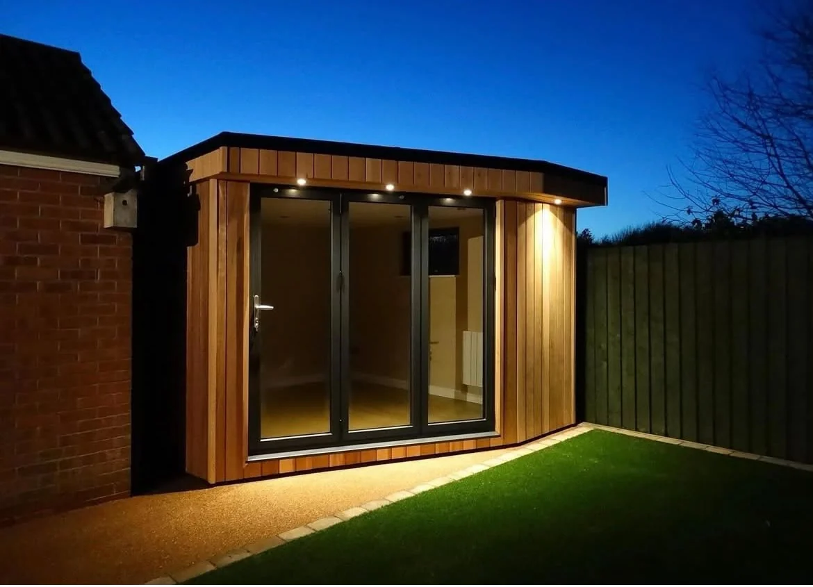 Compact timber-clad garden room with glazed doors and warm exterior lighting at dusk