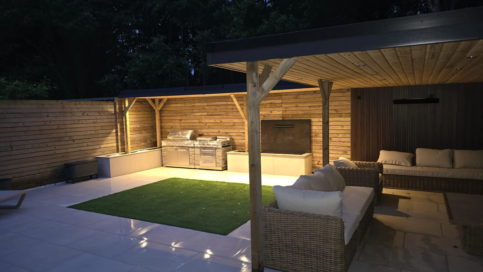 Illuminated timber pergola sheltering an outdoor seating area beside timber screening and a barbecue area